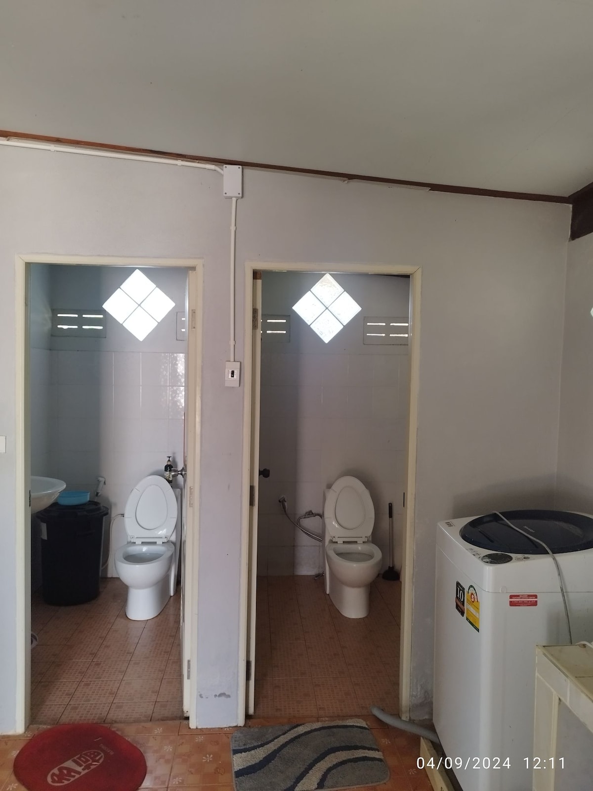 A clean and functional bathroom space is presented, featuring two individual bathroom stalls with white toilets. Each stall is equipped with a small window allowing natural light in. A washing machine is visible, positioned against one wall, adding convenience to the space.