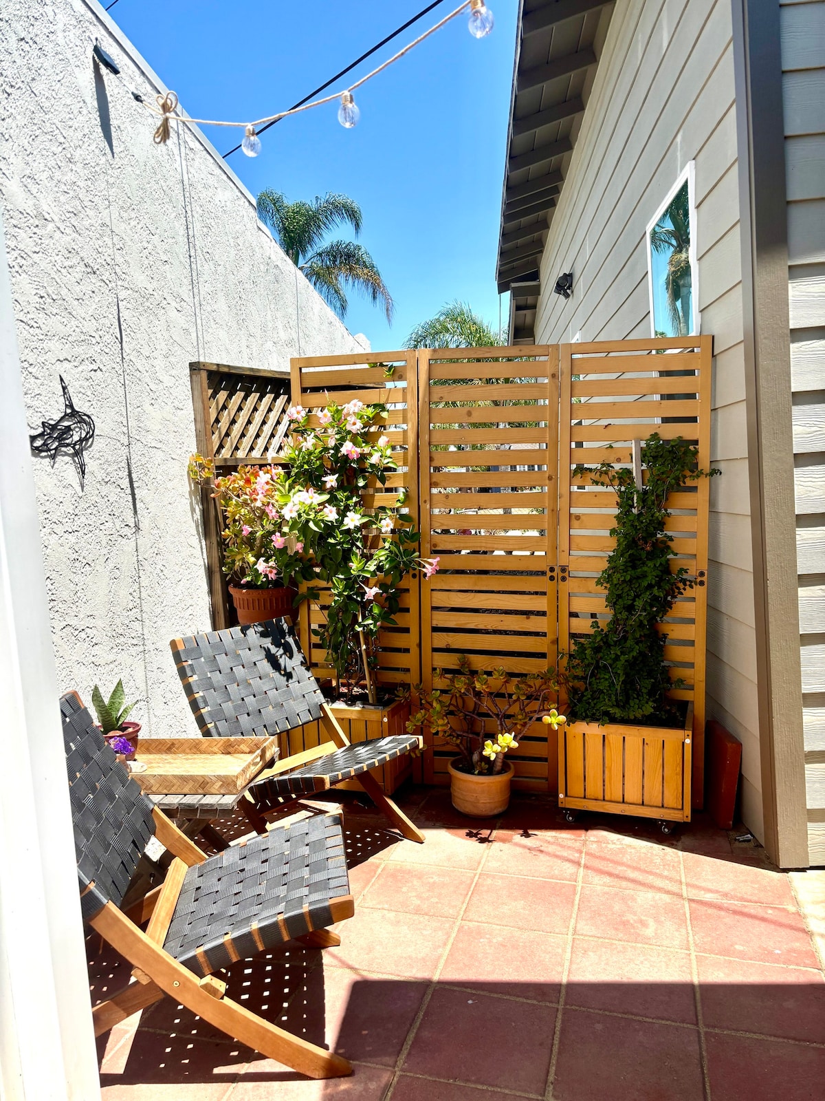 An outdoor seating area features two wooden chairs positioned on a tiled surface. A wooden privacy screen supports greenery, including climbing plants and potted flowers, creating a serene atmosphere. Sunlight illuminates the space, enhancing the natural elements present in the area.