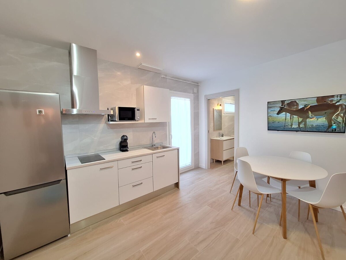 A modern kitchen area is presented, featuring sleek white cabinetry and stainless steel appliances. A round dining table with four chairs sits nearby, and light enters through a glass door leading to an outdoor space. A framed artwork depicting dogs is displayed on the wall.