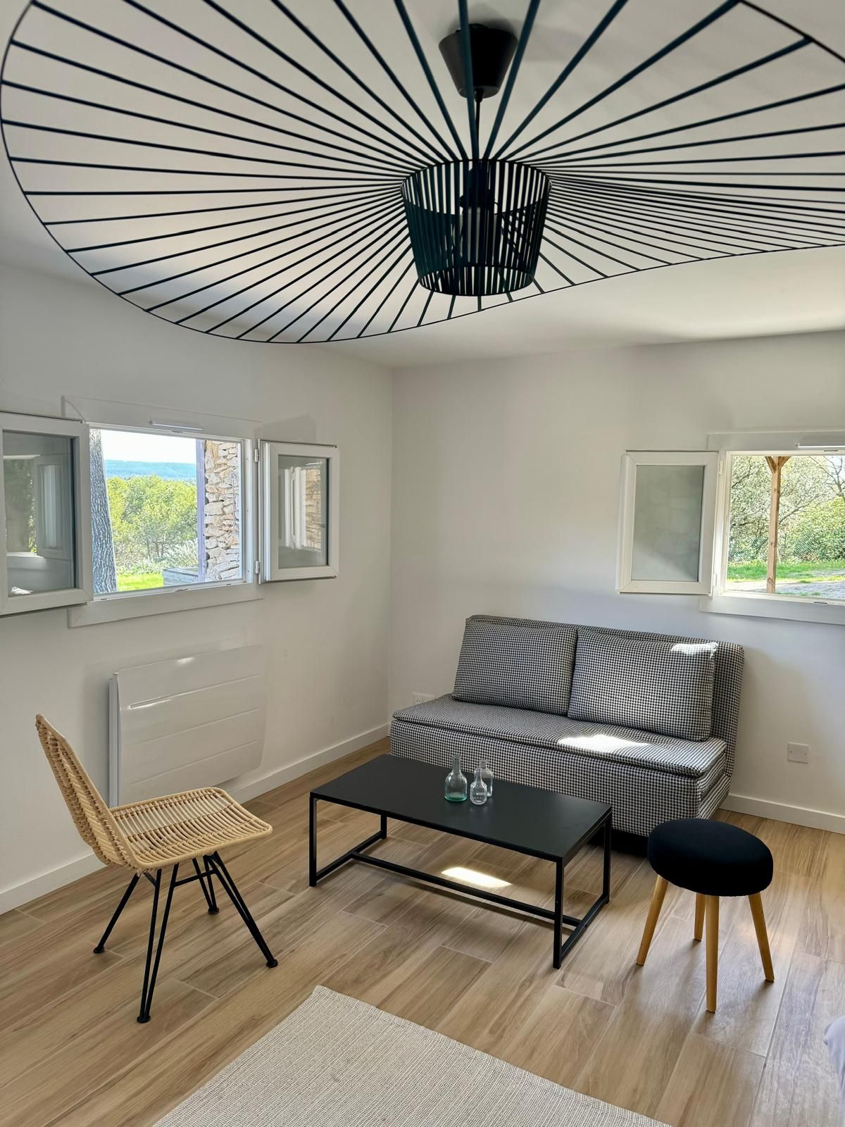 A bright living area features a modern gray sofa accompanied by a black coffee table and a small round stool. Natural light enters through two open windows, highlighting the warm wooden flooring. A woven chair adds texture to the space, and a distinctive light fixture adorns the ceiling.