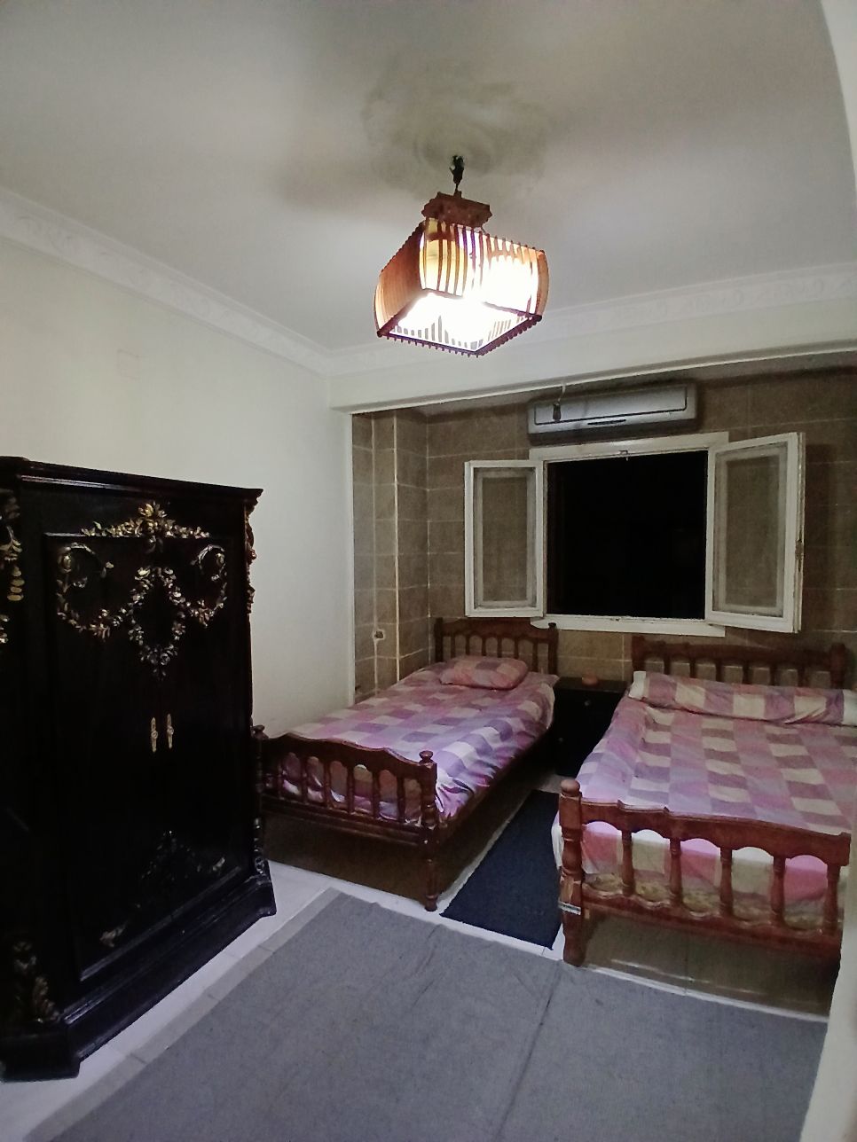 A room is furnished with two twin beds, each made with patterned bedding. An antique wardrobe stands against one wall, and a window with multiple panes allows natural light to enter. An air conditioning unit is mounted above the window.