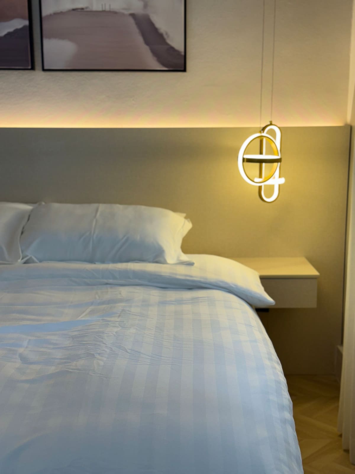 A neutral-toned bedroom features a spacious bed with crisp white linens. A minimalist pendant light hangs above a bedside table, creating subtle illumination. A soft glow highlights the headboard, while abstract art pieces adorn the wall in the background.