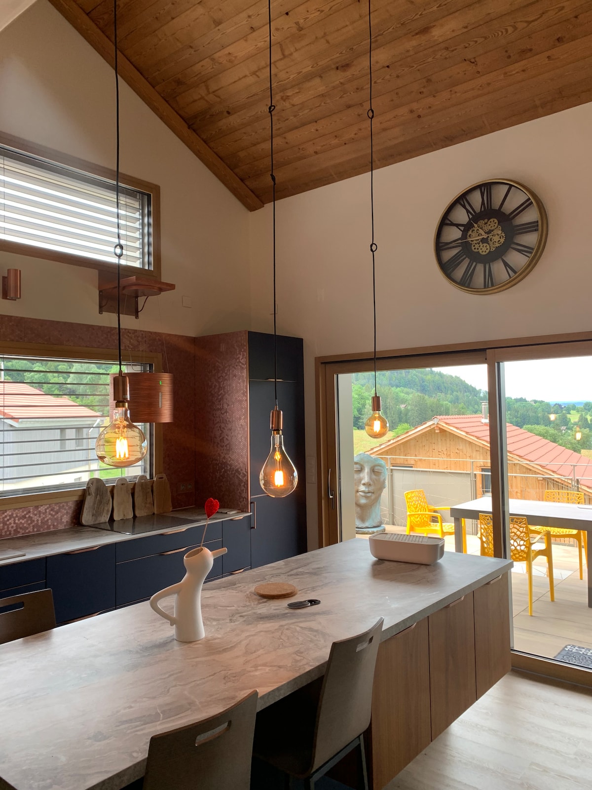 A modern kitchen features a wooden ceiling and is illuminated by stylish pendant lights. A large island with a marble countertop serves as a central space. Large glass doors open to a scenic view, with outdoor seating visible on the terrace beyond.
