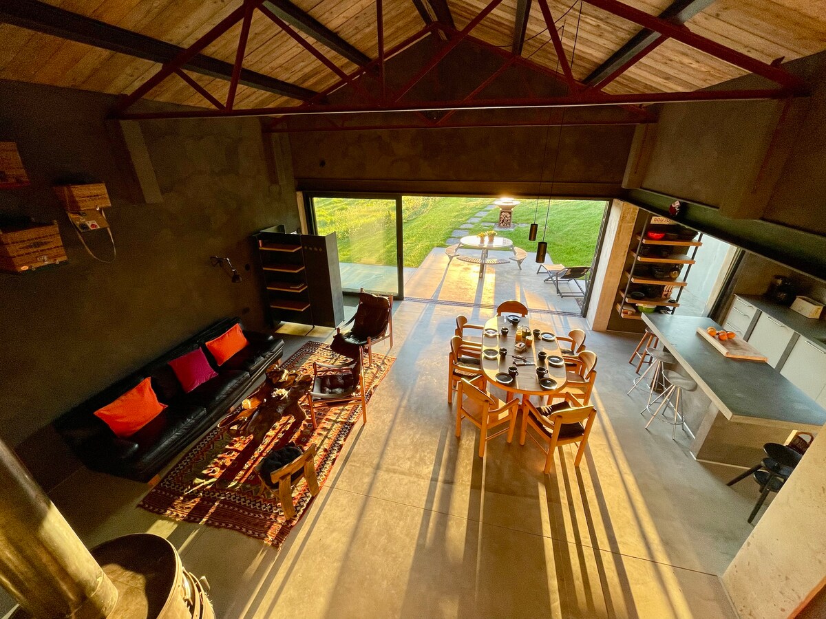 A spacious living area features a mix of modern design and rustic elements. A large table is set for dining, while a black sofa adorned with colorful cushions offers a cozy seating option. The expansive windows invite natural light and provide a view of the green surroundings.