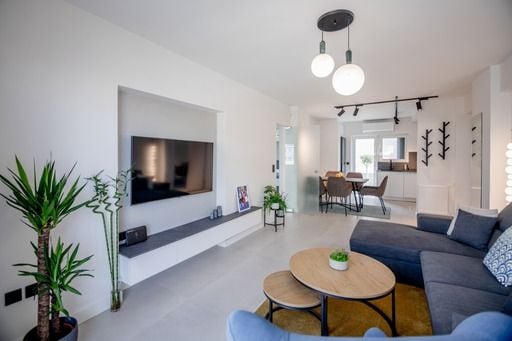 A modern living area features a spacious sectional sofa, two round coffee tables, and a large wall-mounted television. Plants are positioned in the corners, adding a touch of greenery. Natural light fills the room through large glass doors that lead to an outdoor space.