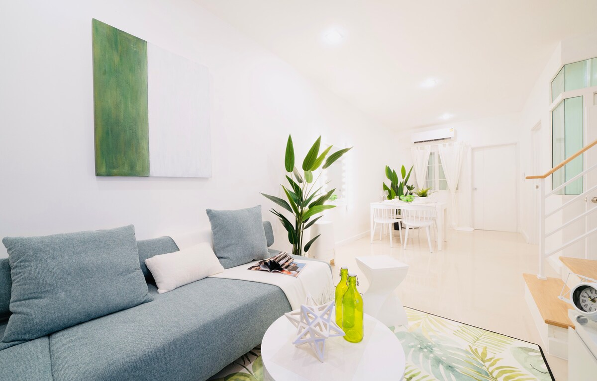 A light-filled living space features a sectional sofa adorned with decorative pillows. A minimalist white coffee table is accompanied by a vibrant green plant and decorative items. The room includes a dining area with a white table and chairs visible in the background, accentuated by large windows.