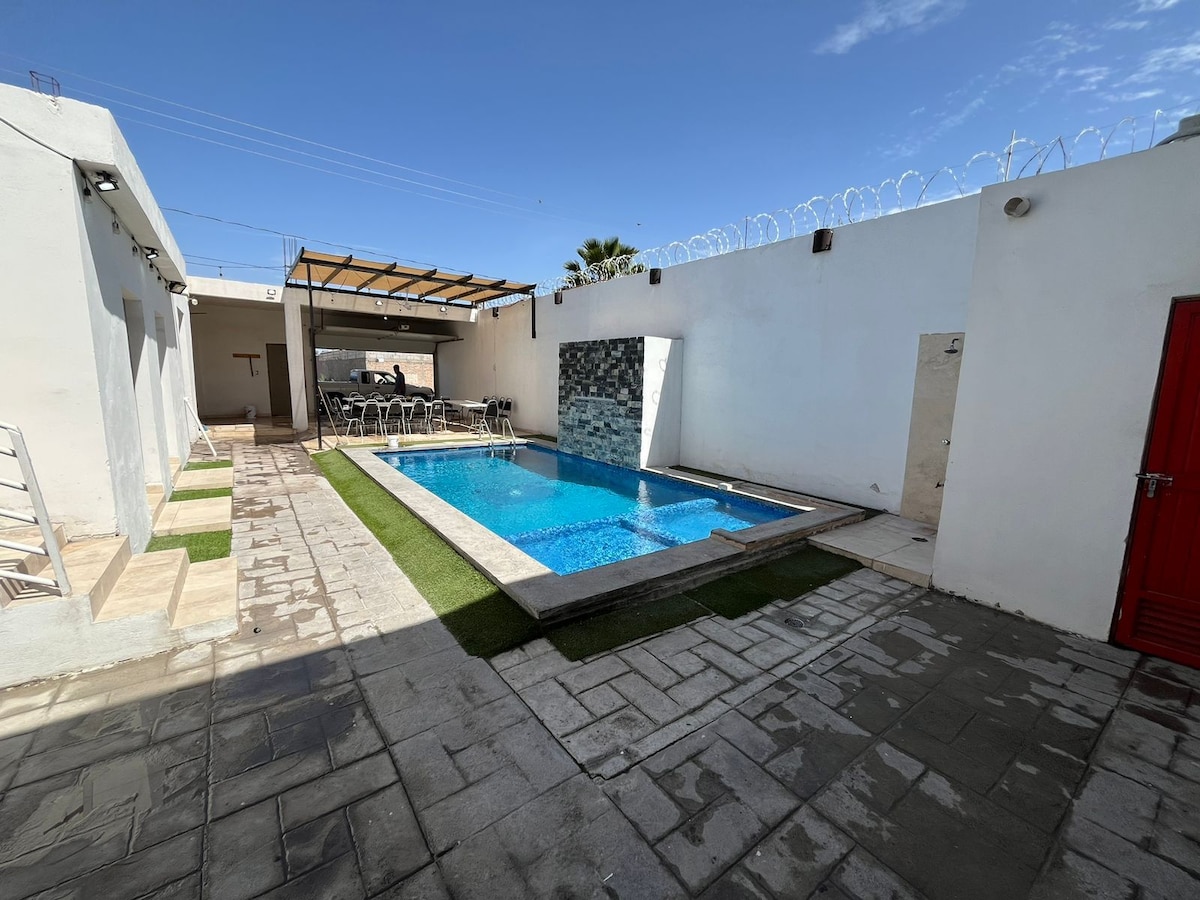 A tranquil outdoor space features a crystal-clear pool surrounded by neatly arranged stones and artificial grass. Chairs are positioned around the pool, ideal for relaxation. High walls provide privacy, with a textured stone wall adding visual interest on one side.