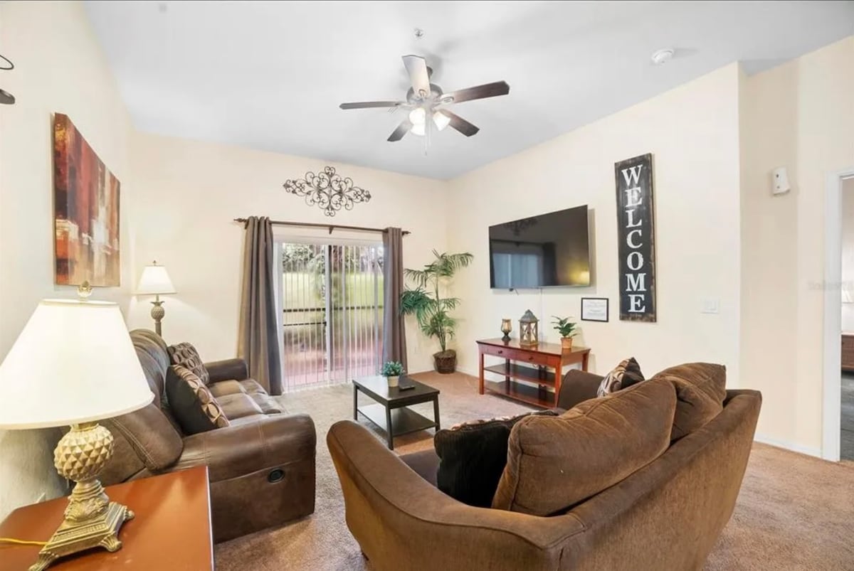 A spacious living area features a comfortable brown couch and armchair, arranged around a coffee table. A ceiling fan hangs above, and a large flat-screen TV is mounted on the wall. Natural light enters through sliding glass doors, which lead to an outdoor space.