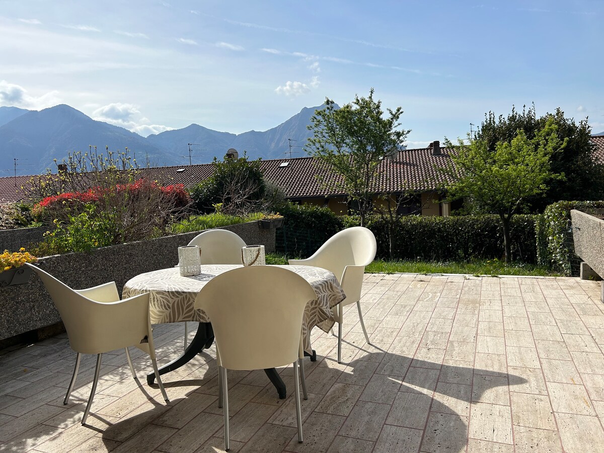 A spacious terrace is showcased, featuring a round table surrounded by four white chairs. Lush greenery and colorful flowers border the area, with distant mountain views framing the sky. Sunlight illuminates the outdoor space, enhancing the serene outdoor setting.