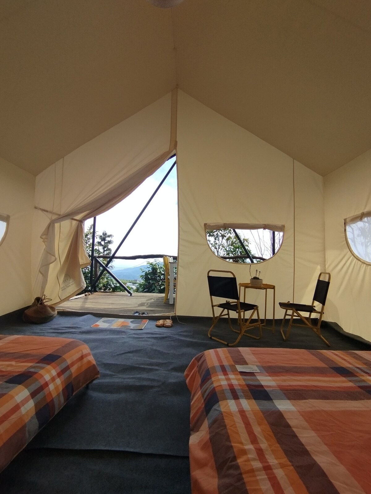 An airy tent interior is presented with two spacious beds featuring plaid bedding. A small table and two folding chairs are positioned near the entrance, which opens to a scenic view framed by large windows, creating an inviting environment that connects with nature.