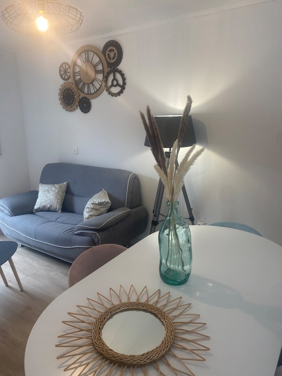 A cozy living area is featured, with a gray sofa adorned with decorative pillows. A modern floor lamp provides soft lighting, while an arrangement of gears decorates the wall. A dining table is set with pastel chairs, accompanied by a decorative mirror and a vase of dried plants.