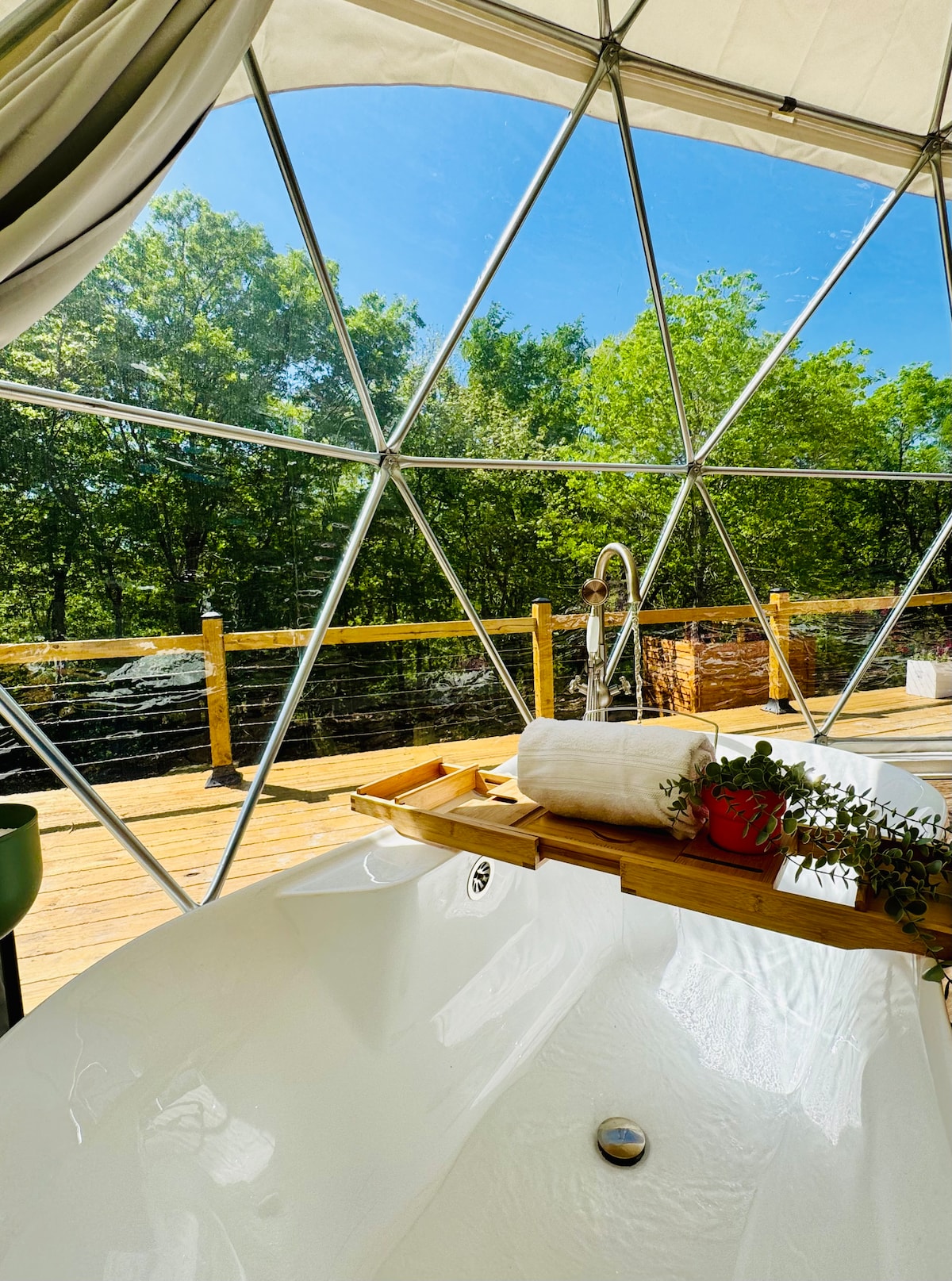 A vintage clawfoot tub is positioned beside a panoramic window, offering a view of lush greenery outside. A wooden bath tray is placed across the tub, adorned with a small potted plant and a rolled towel, inviting moments of relaxation and connection with nature.