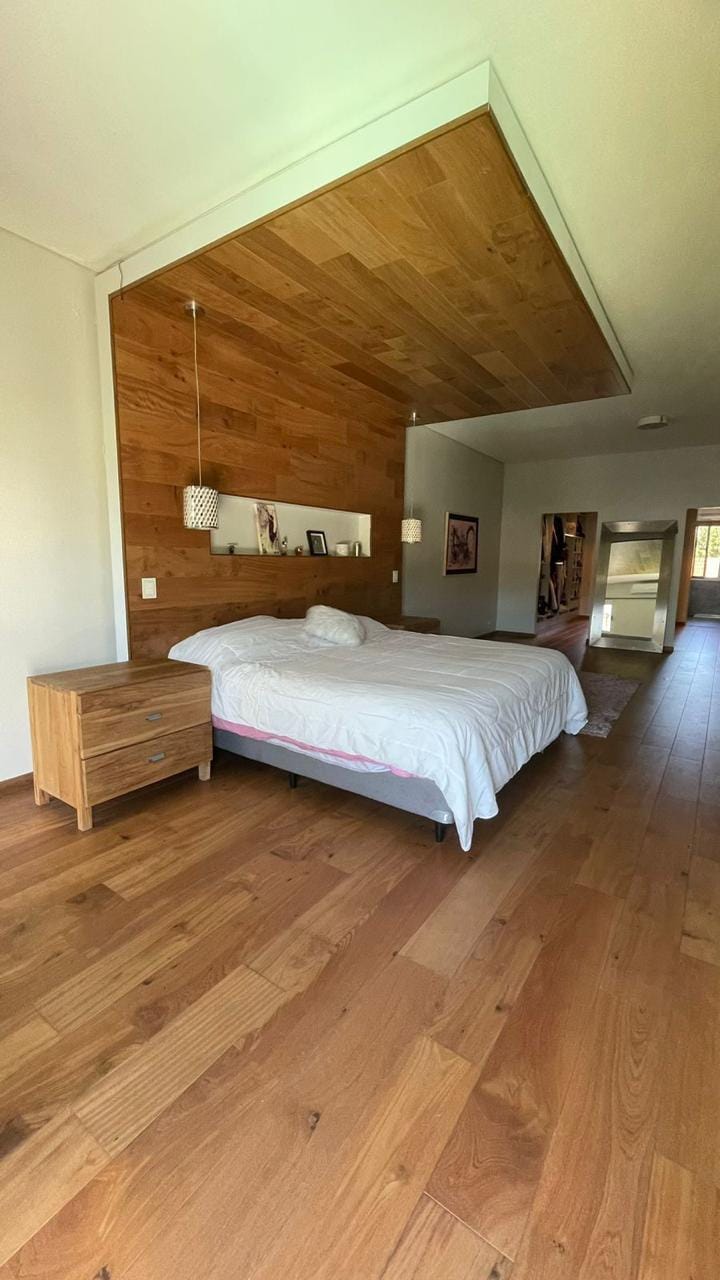 The bedroom features a spacious bed positioned against a stylish wooden accent wall. Soft lighting is provided by pendant fixtures on either side. Warm hardwood flooring adds to the inviting atmosphere, and a doorway leads to an adjoining area, suggesting a well-integrated living space.