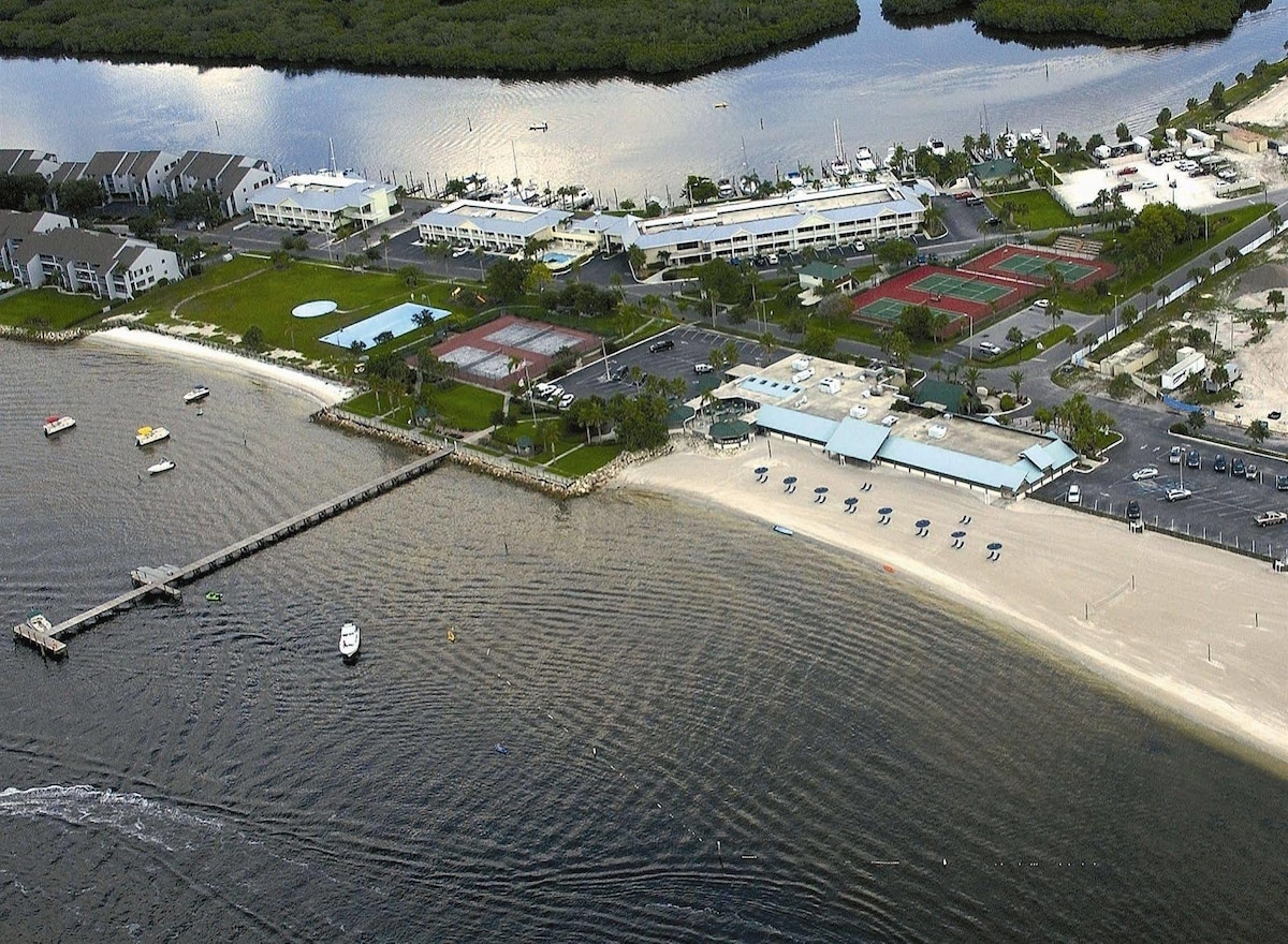 An aerial view illustrates the sandy beach and calm waters leading to the shore. The image captures the docks and various recreational facilities, including tennis courts and a swimming pool, set against the backdrop of lush greenery and nearby buildings.