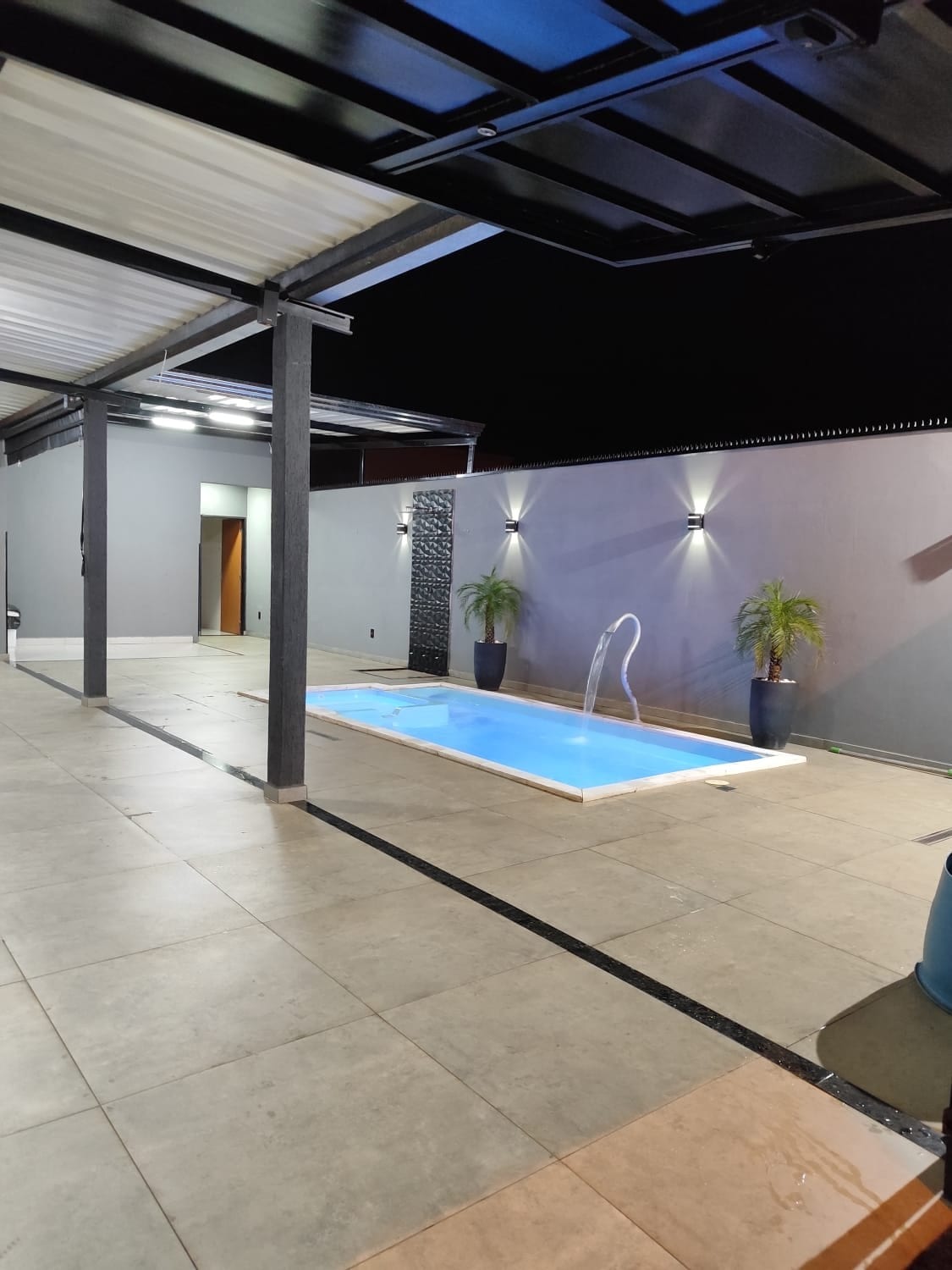 An inviting outdoor area features a modest pool with a gentle water feature, surrounded by sleek tile flooring. Soft lighting highlights the walls, and two potted plants add a touch of greenery to the space. A covered seating area is visible in the background.