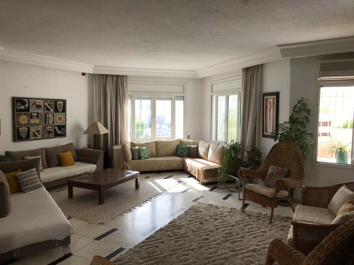 A spacious living area features light-colored sofas arranged around a central wooden coffee table, complemented by a textured area rug. Natural light filters through large windows, highlighting potted plants and cozy wicker chairs. Soft curtains frame the windows, creating a warm and inviting space.