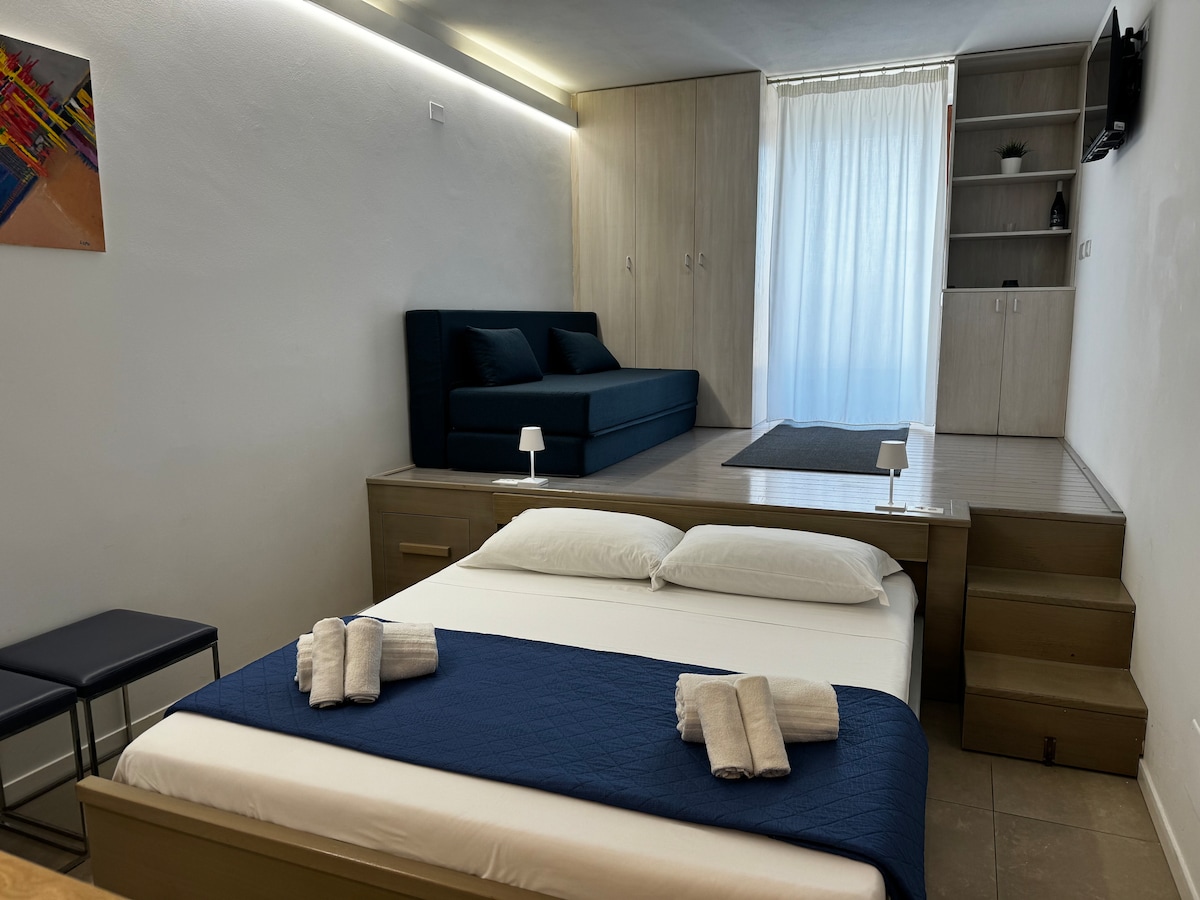 A modern living space features a pull-down bed with crisp white linens, complemented by a navy throw blanket. A comfortable sofa bed is positioned behind, while natural light filters in through a sheer curtain. Additional storage is provided with built-in cabinetry along the wall.