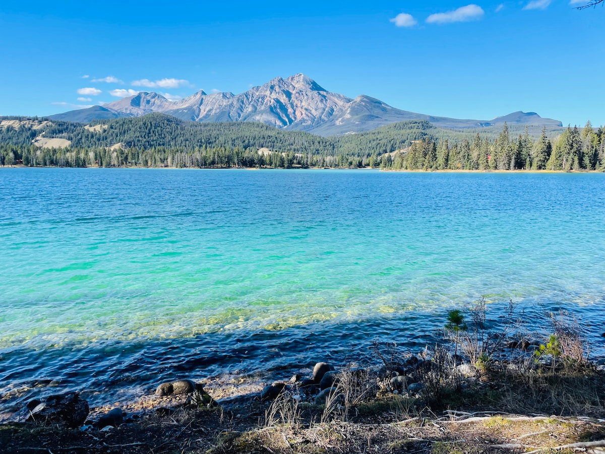 A serene lake is framed by a lush forest and mountains in the background. The water displays a gradient from deep blue to vibrant turquoise, reflecting the clear blue sky above. Pebbles and rocks line the shore, adding texture to the tranquil landscape.