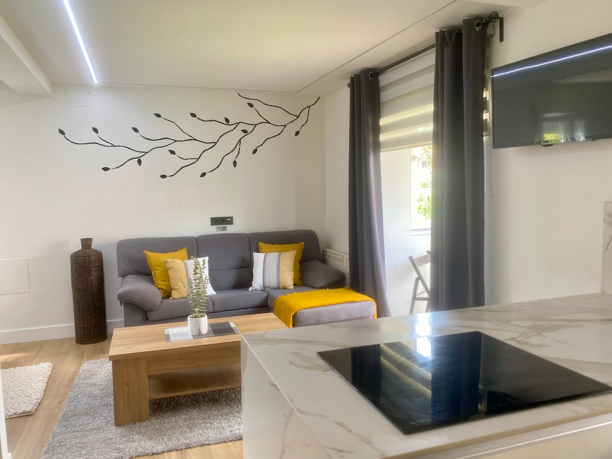 A comfortable living area is showcased with a gray sofa adorned with yellow and white pillows. A wooden coffee table is positioned in front, complemented by a decorative plant. Natural light filters through the window treatments, and a wall-mounted TV is visible. A small dining area can be seen in the background.