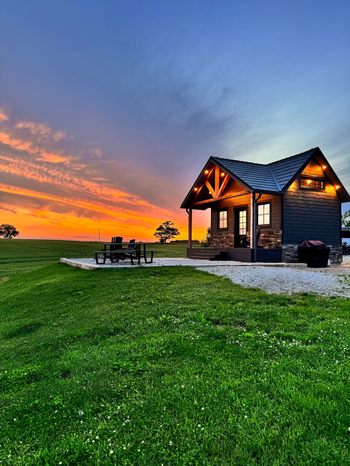 An inviting tiny house is set against a vibrant sunset, featuring warm lighting that highlights its architectural details. A patio area with outdoor seating is visible, surrounded by lush green grass, creating a peaceful outdoor living space.