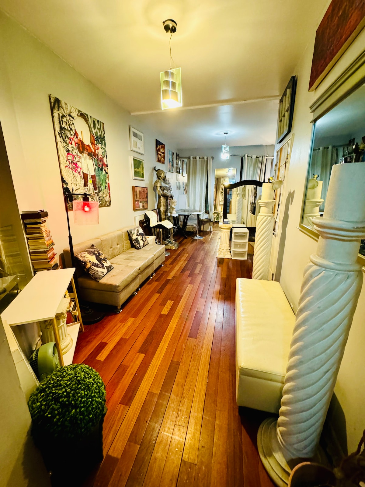 A narrow hallway showcases polished wooden floors, flanked by vibrant artworks on the walls. A plush sofa is positioned next to a tall mirrored cabinet, while a whimsical stature adds character. Soft lighting from pendant fixtures illuminates the space, creating a welcoming environment.