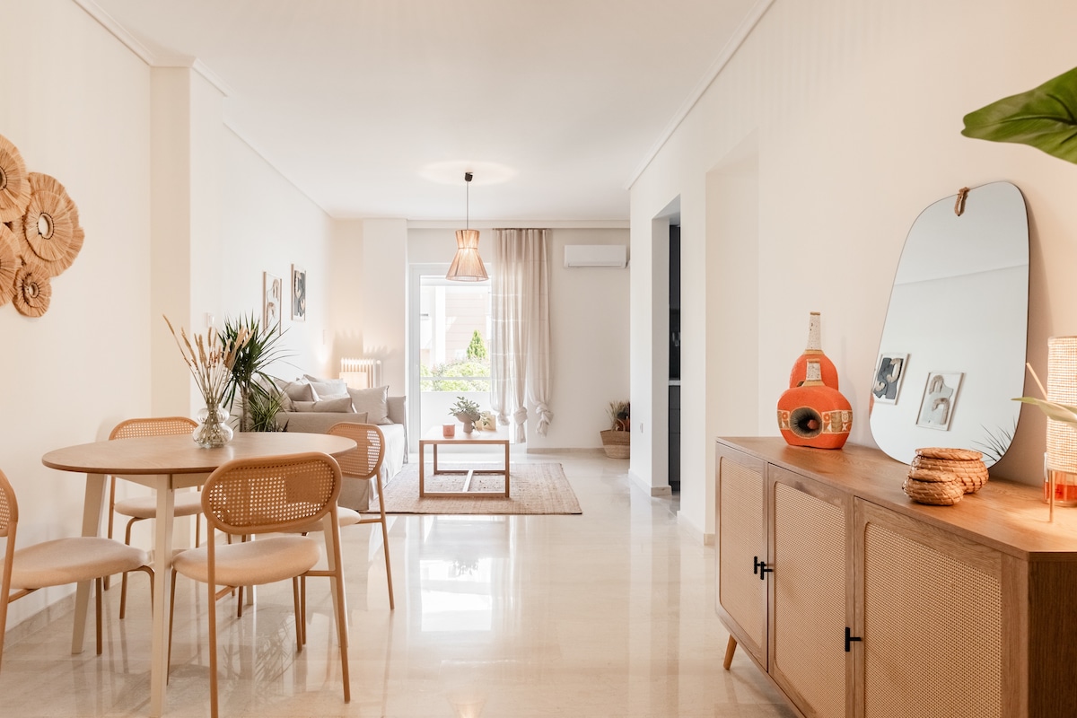 A modern and open living area features a dining table surrounded by four chairs, a stylish sideboard, and a comfortable sofa. Natural light pours in through large windows, illuminating light-coloured walls and minimalistic decor. A neutral rug lies on the polished floor, creating a welcoming ambiance.