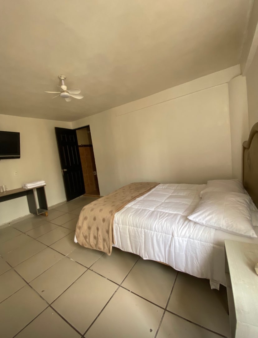 A private sleeping area features a spacious bed with a neutral-colored blanket, against a light-colored wall. A ceiling fan provides airflow, while a television is mounted for entertainment. The room is complemented by tile flooring and a dark wooden door leading to additional space.