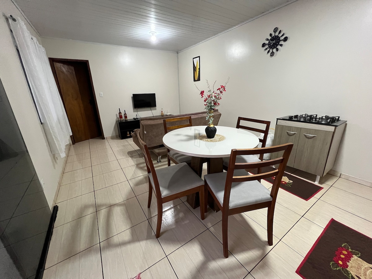 A bright dining area features a round table surrounded by four wooden chairs. A small TV is mounted on the wall, and a kitchenette is visible with a gas stove and cabinetry. Soft lighting enhances the neutral-colored walls.
