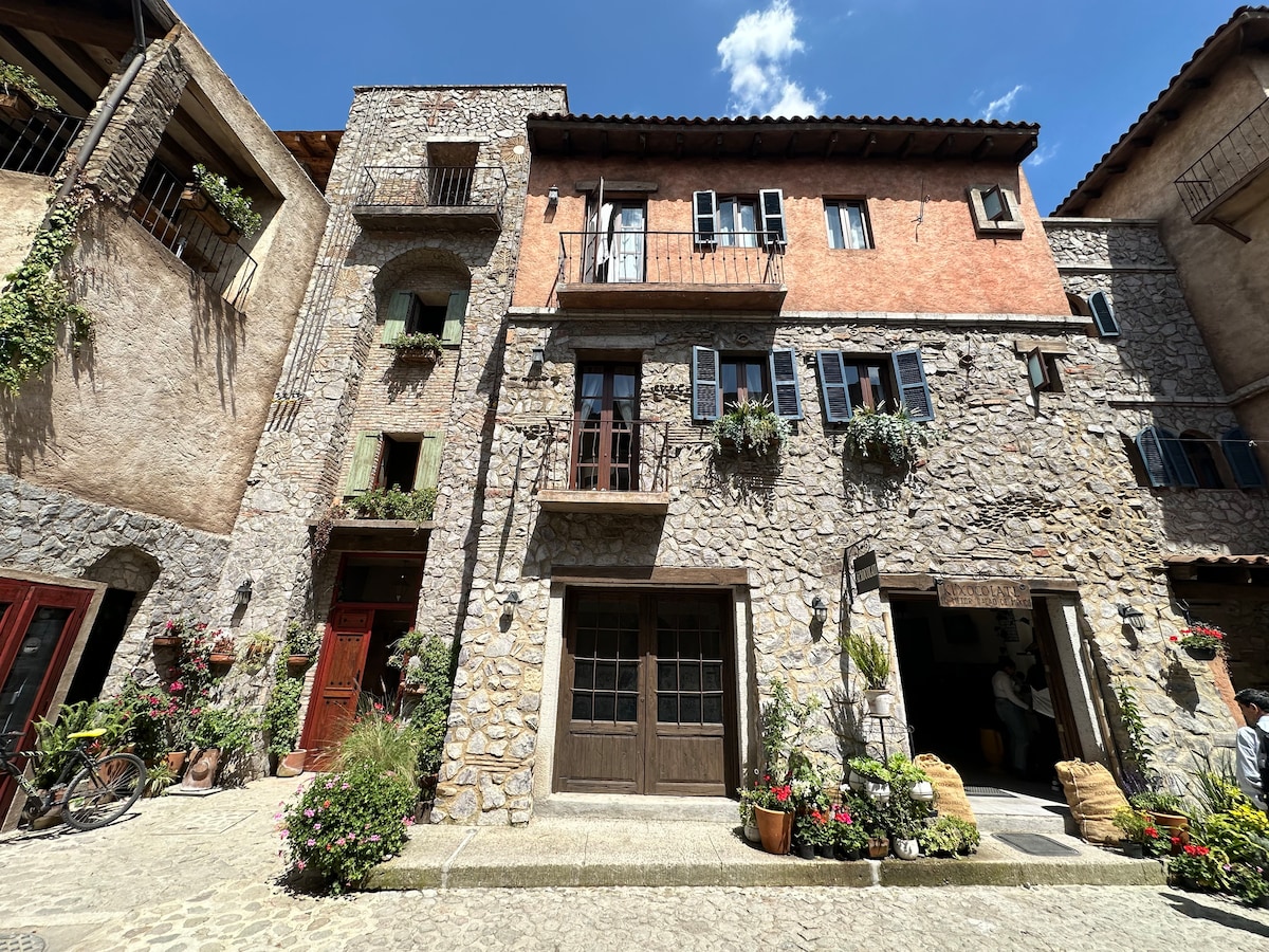 A charming stone building with a mix of rustic and modern elements is displayed. Flowering plants adorn the balconies, and narrow wooden doors invite entry. A cobblestone path leads to the entrance, with various decorative elements enhancing the inviting exterior.
