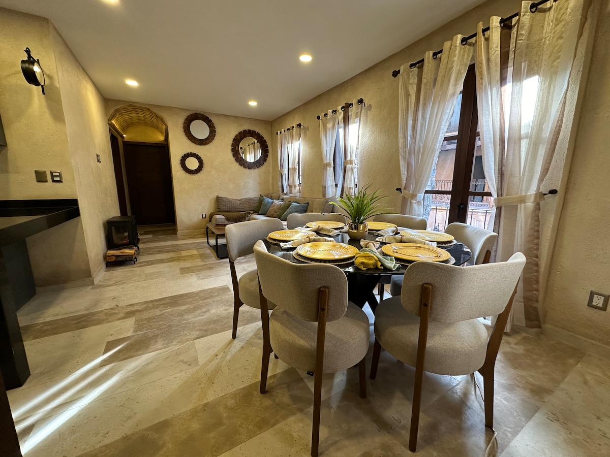 A dining area is presented with a round table set for six, accompanied by comfortable upholstered chairs. Natural light enters through sheer curtains, illuminating the space. Decorative mirrors adorn the walls, while a cozy seating area is visible in the background.