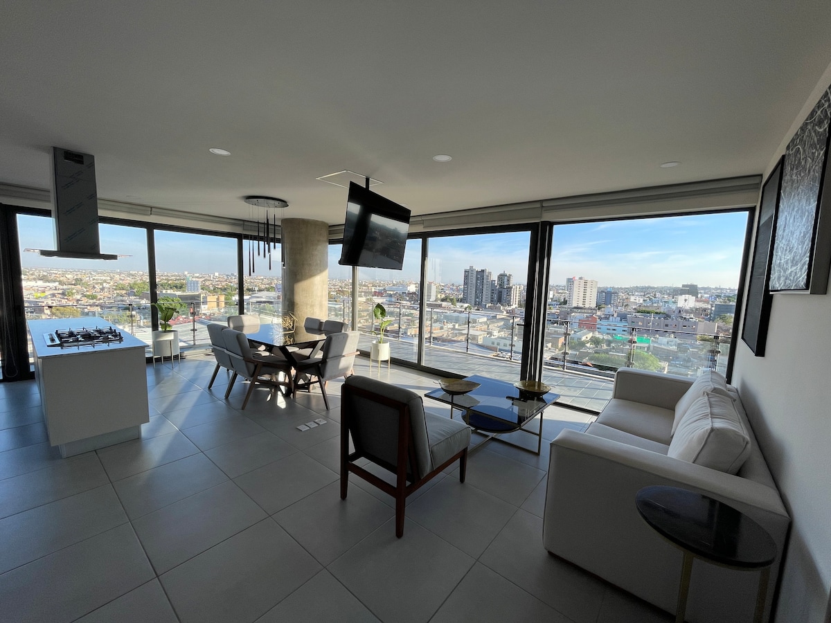 A modern living space is highlighted by large floor-to-ceiling windows, offering expansive views of the city. A comfortable sofa and dark wooden accent chairs surround a sleek glass coffee table, while a dining table for six is positioned nearby. Bright natural light fills the room.