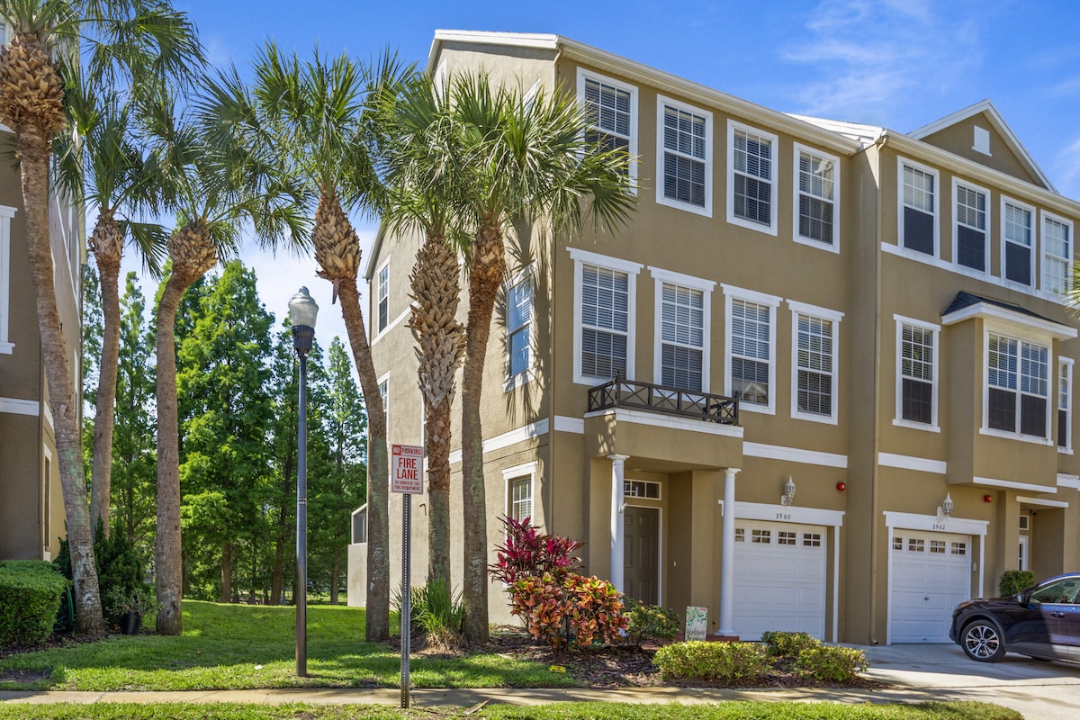 The exterior of the townhome is presented, showcasing a corner unit adorned with multiple large windows. Lush palm trees and vibrant landscaping frame the entrance. A one-car garage is visible, along with a well-kept pathway leading to the front door.
