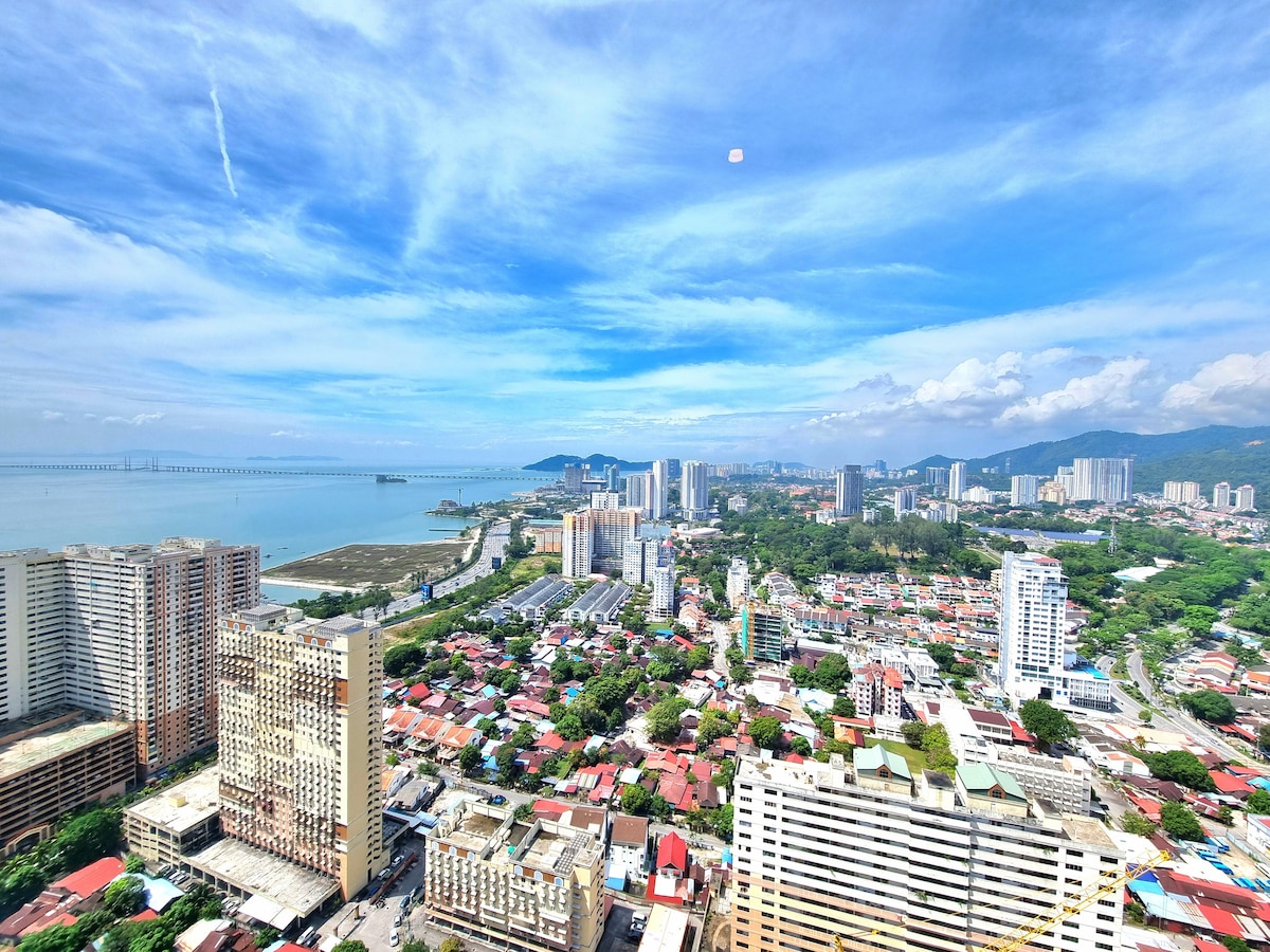 An expansive view from a high vantage point showcases a mix of urban and coastal landscapes. The ocean glistens under a clear blue sky, while vibrant cityscapes and greenery complement the scene. The Penang Bridge is visible in the distance, connecting land and sea.