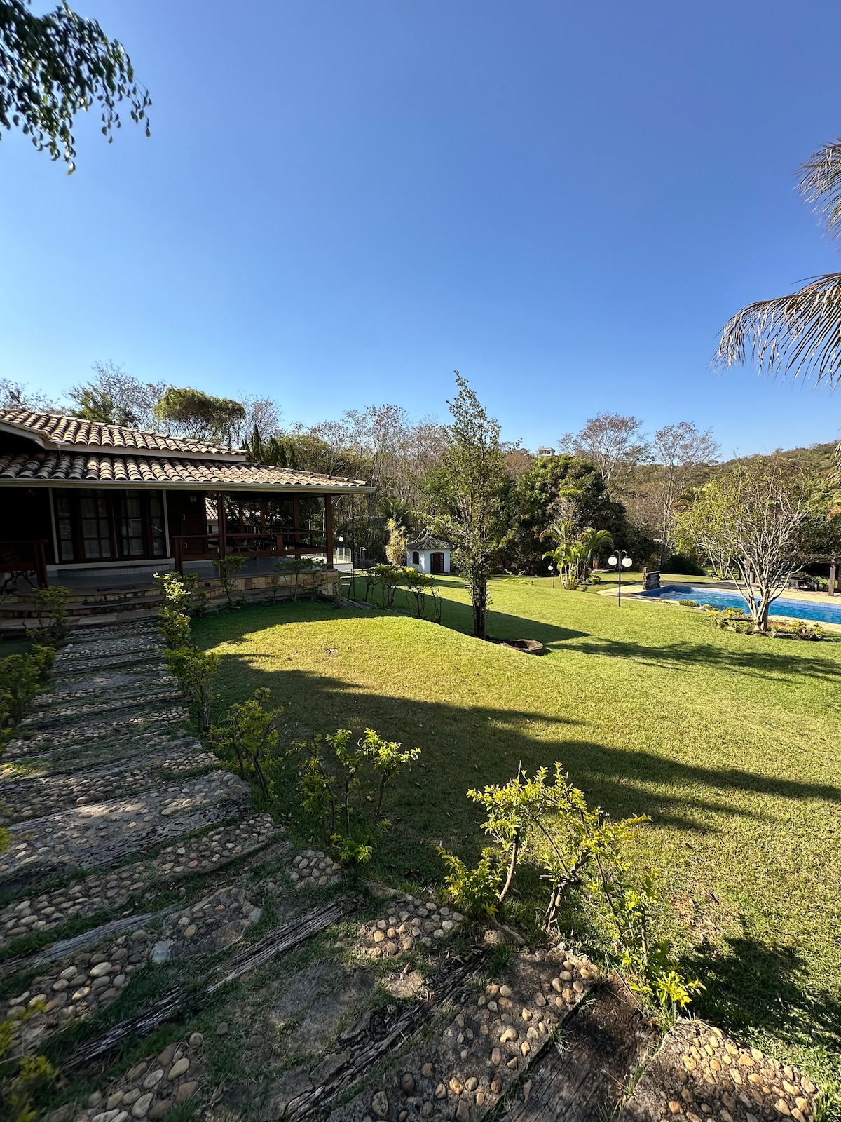 A scenic outdoor view showcases a well-maintained lawn bordered by neatly arranged stones. The image includes a wooden house with large windows, surrounded by trees and greenery, with a glimpse of a pool in the background under a clear blue sky.