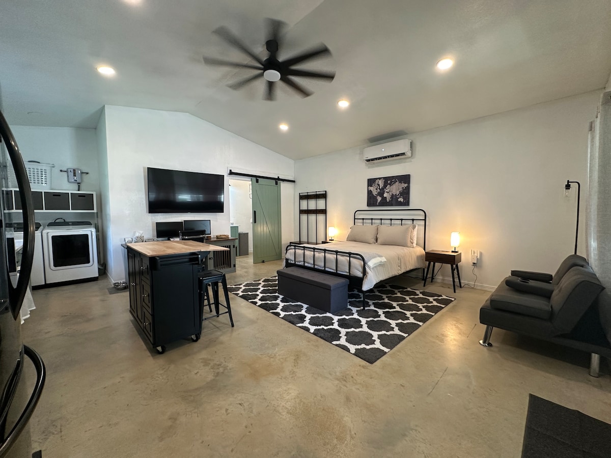An open floor plan studio features a spacious bed with neutral bedding, complemented by a patterned area rug. A dining table with chairs is positioned nearby, along with a modern sofa. A ceiling fan and wall-mounted TV add to the airy ambiance.