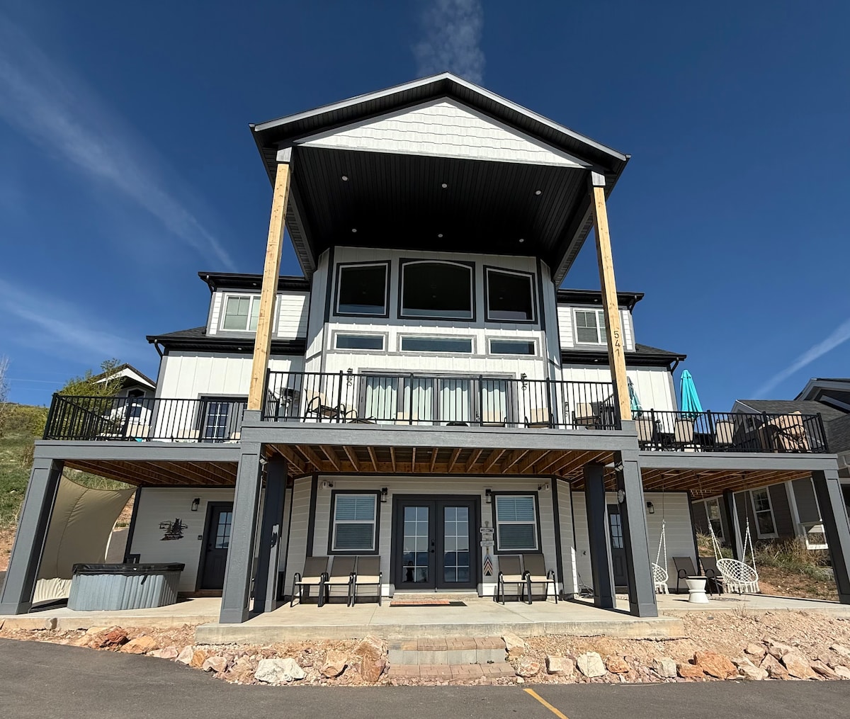 The exterior of Raspberry Sunrise Vacation Rental displays a modern design with multiple levels and expansive balconies. Large glass doors invite natural light, while the deck furniture and an umbrella offer outdoor seating options. A hot tub is visible on the lower patio.