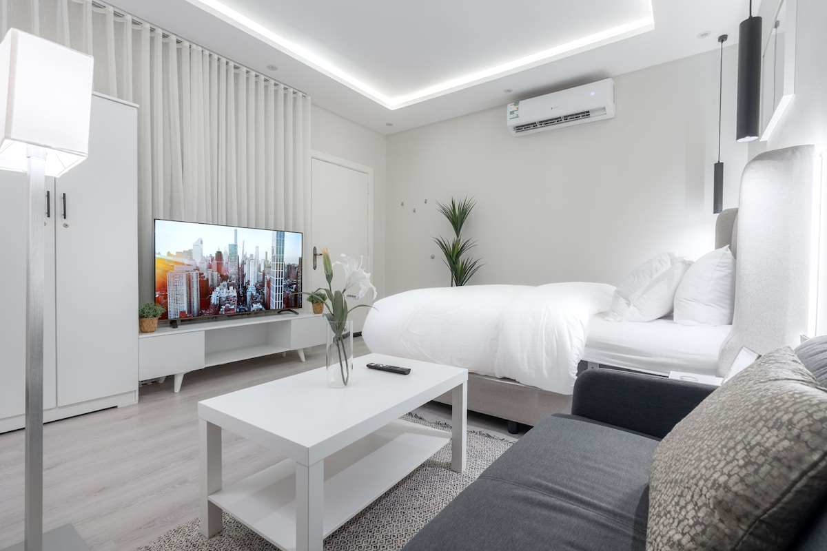 A modern studio features a spacious bed with white bedding, positioned near a contemporary entertainment center with a large TV. A minimalist coffee table sits in front of a dark sofa, and greenery adds a fresh touch to the light-filled space.
