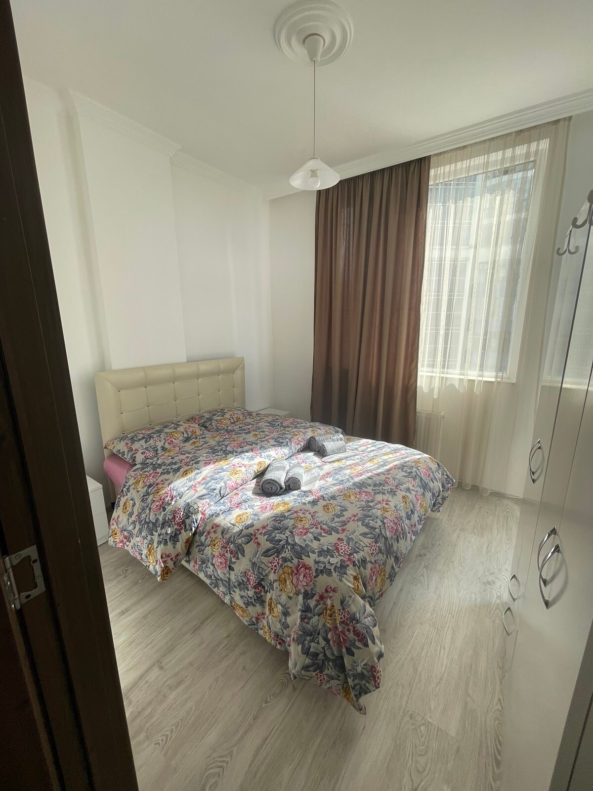 A well-lit bedroom features a spacious bed adorned with a floral patterned blanket. Natural light enters through a large window, complemented by sheer curtains that softly diffuse the light. A cozy atmosphere is created by light-colored walls and earthy wooden flooring.