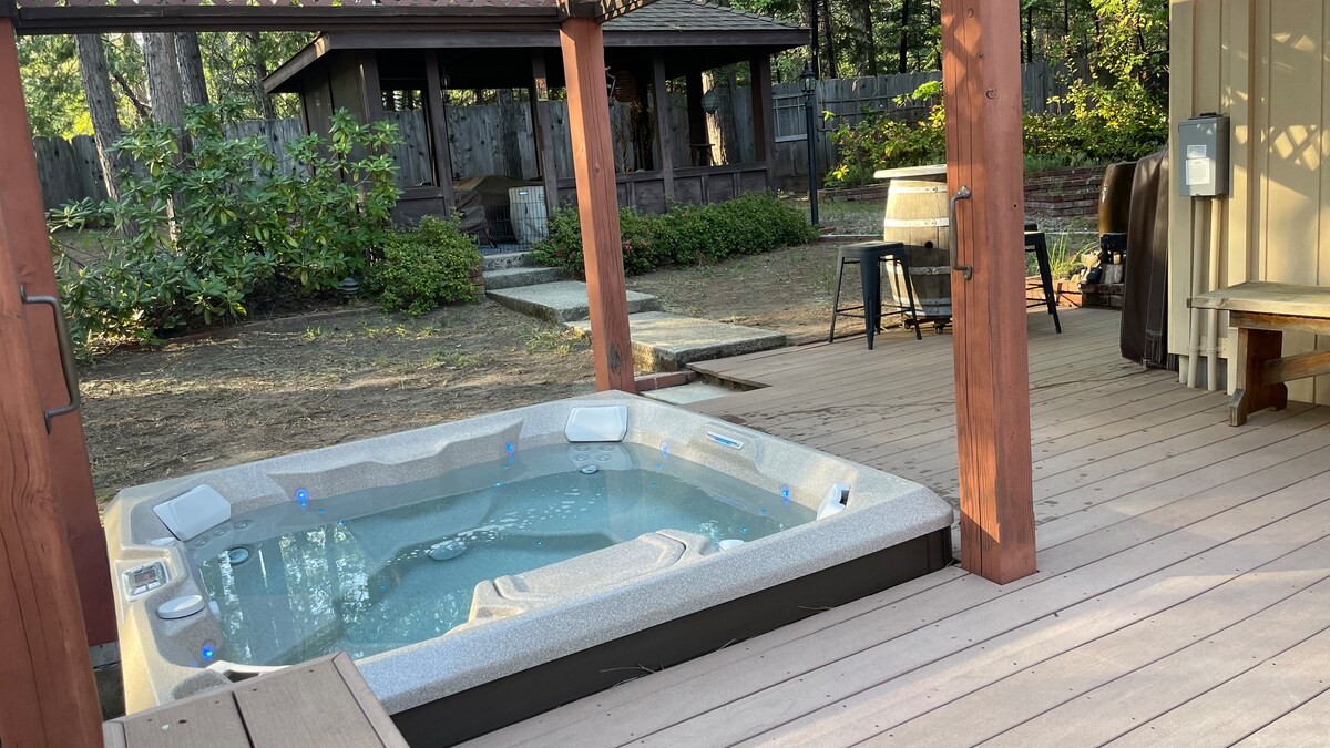 A hot tub is positioned on a wooden deck, surrounded by a serene outdoor setting. In the background, a gazebo is visible, amid trees and a cleanly landscaped yard. Relaxation spaces are presented, creating an inviting atmosphere for outdoor enjoyment.