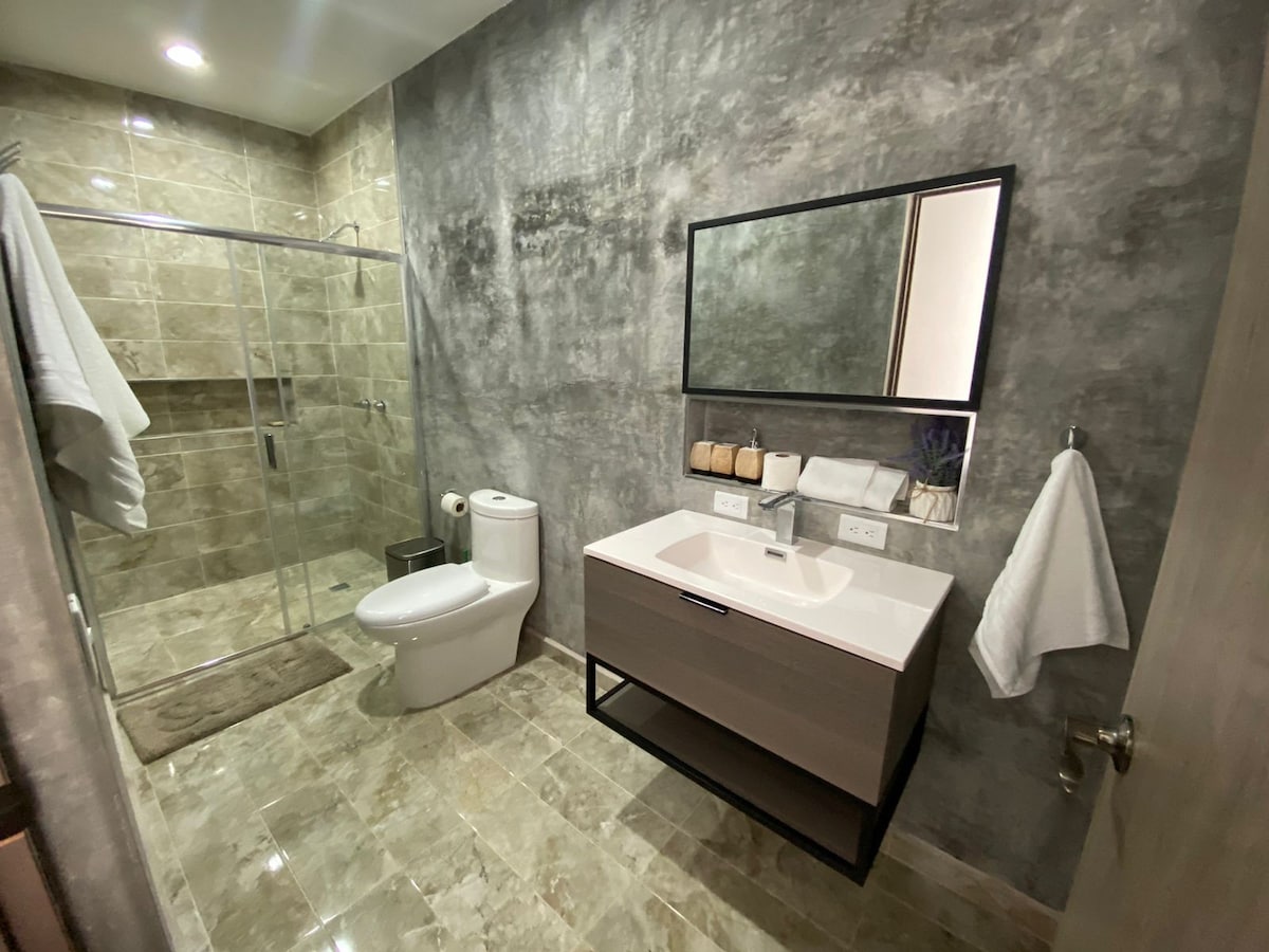 An elegantly designed bathroom features modern fixtures and a spacious glass shower. A large mirror above the sink reflects the neutral-toned walls and floor. Clean towels are neatly arranged, and essential toiletries are available on a nearby shelf.