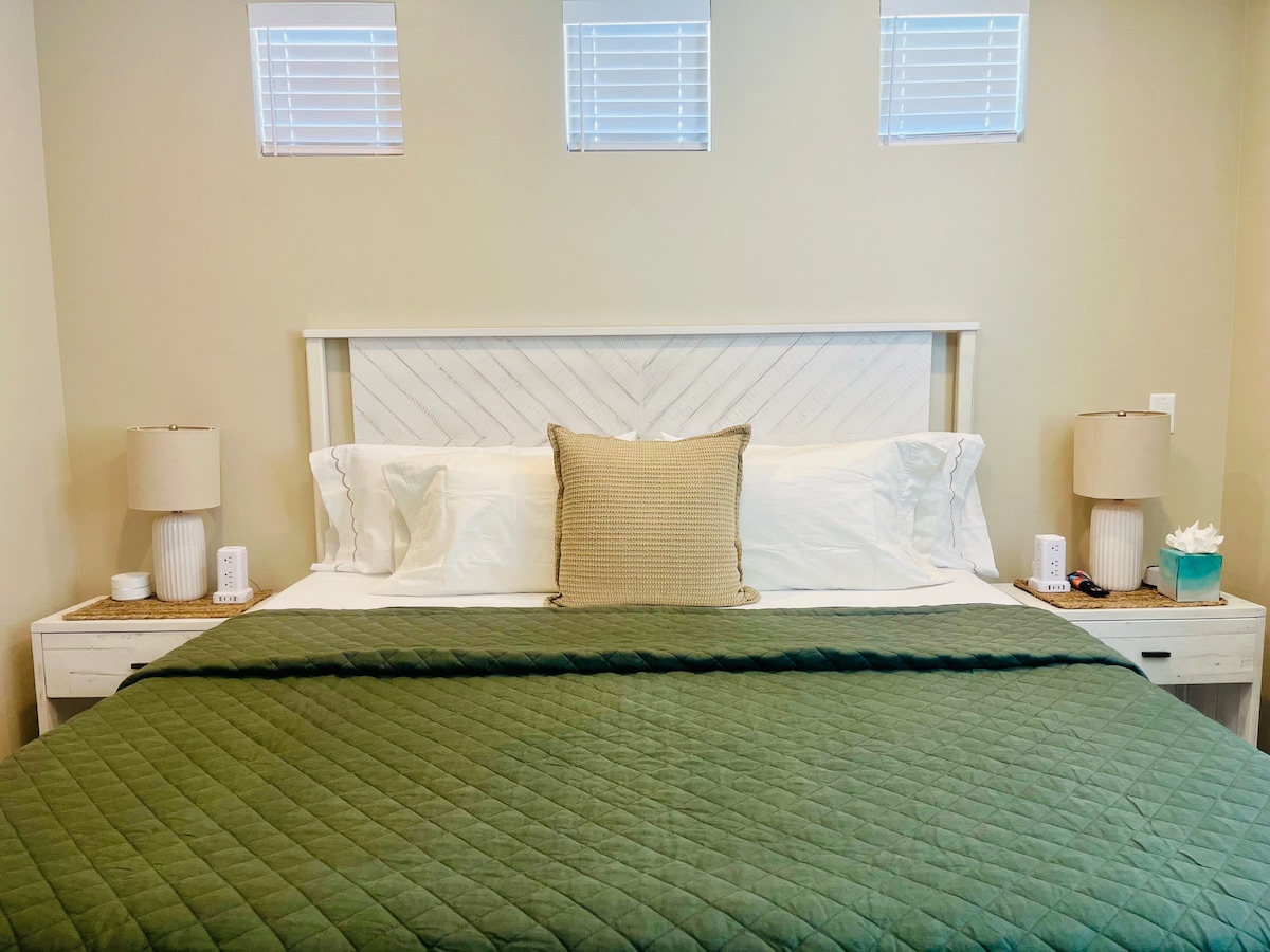 A spacious bed is adorned with a green quilt and decorative pillows. Nightstands with lamps are placed on either side. Three small windows above allow natural light to softly illuminate the room, against light beige walls creating a calm environment.