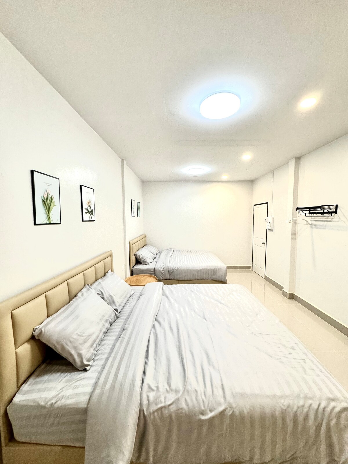 A well-lit sleeping area showcases two spacious beds with neutral-colored linens. Light enters through a circular ceiling fixture, while framed botanical prints adorn the white walls. A simple wooden nightstand complements the decor, creating a calm and welcoming environment.