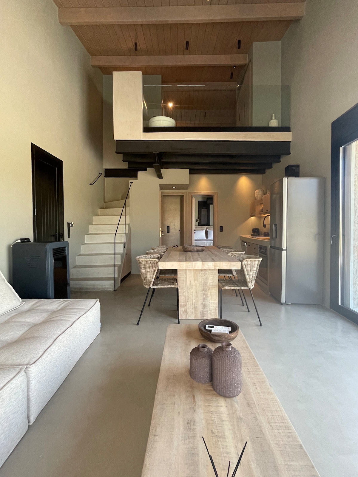An open living area features a light-colored sofa and a wooden dining table surrounded by woven chairs. A staircase leads to an upper level, while a large window allows ample natural light. Kitchen appliances are arranged in a sleek, modern layout.