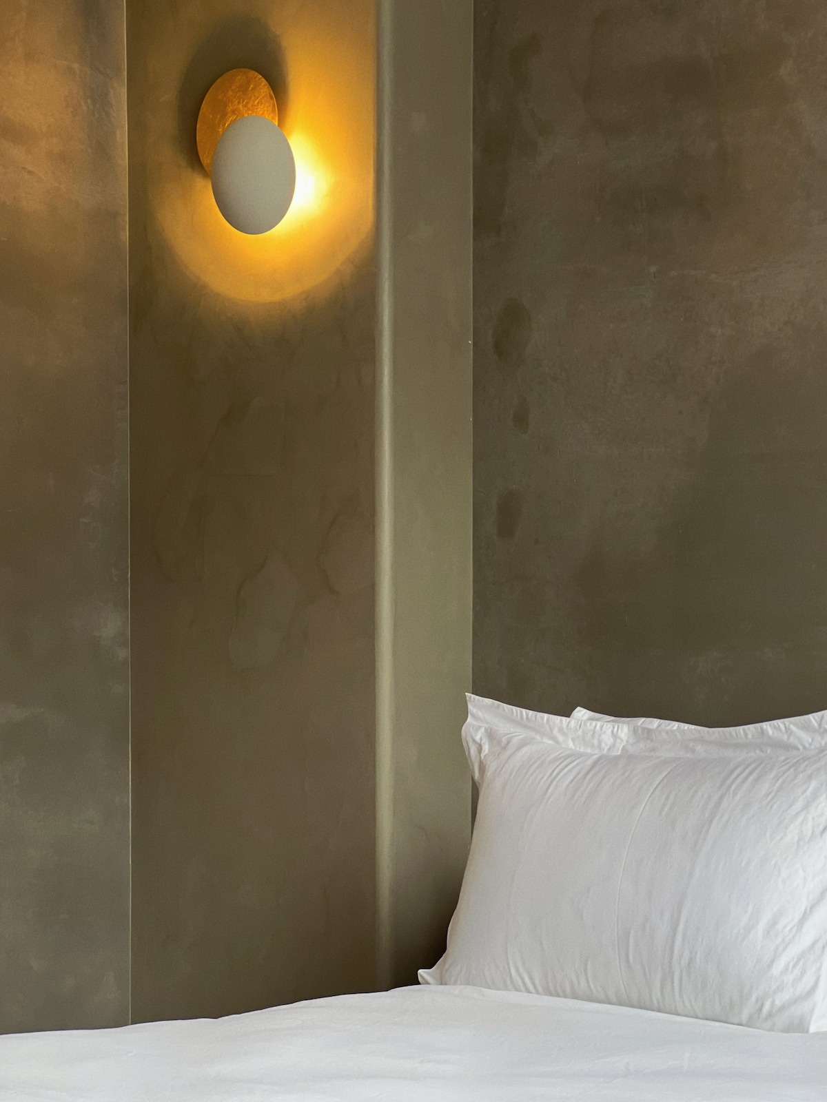 A softly illuminated corner features a spacious bed with crisp white bedding. A wall-mounted light emits a warm glow, highlighting the textural concrete walls, creating a calming and serene environment.