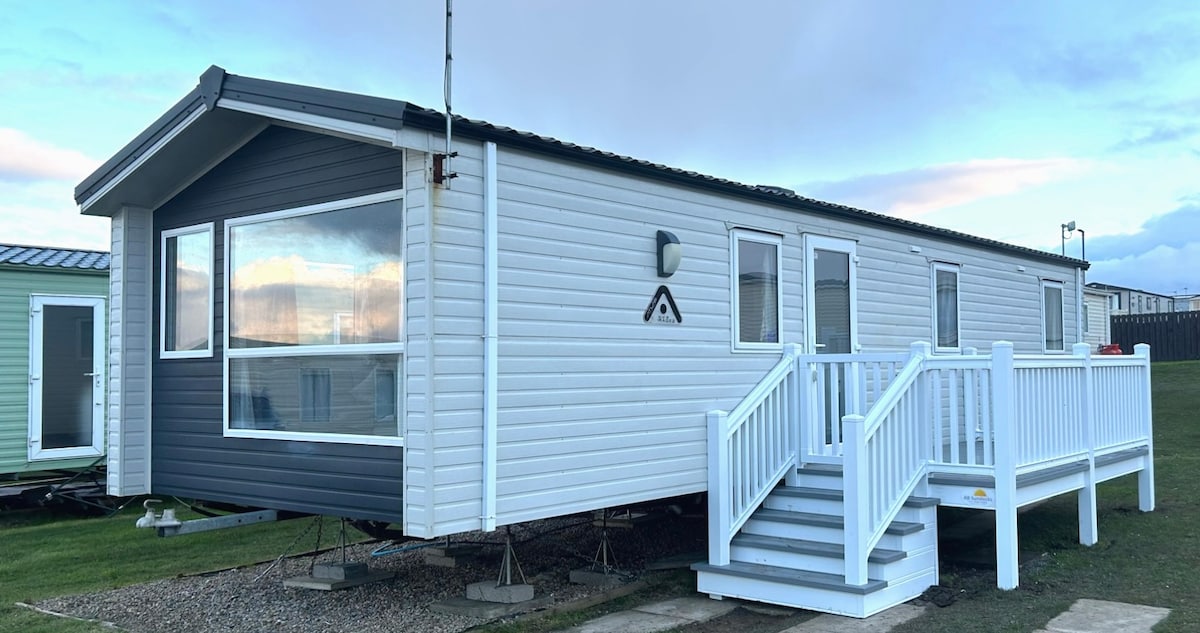 A modern static caravan is displayed from the exterior, featuring a blend of white and dark grey paneling. The structure is raised on stilts, with a sturdy white railing and steps leading up to the entrance. Large windows allow for ample natural light.