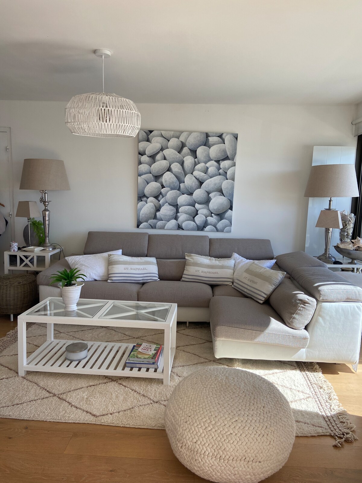 A light-filled living area features a comfortable sectional sofa and a glass-top coffee table resting on a textured area rug. A decorative piece with smooth stones adorns the wall, while stylish lamp shades provide soft lighting, enhancing the room's relaxed atmosphere.