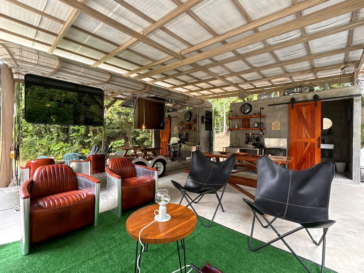 An open-air seating area features stylish leather chairs and modern outdoor furniture, inviting relaxation. A wooden table holds a decorative lamp, and a large screen is mounted on the wall, merging comfort with entertainment. Lush greenery surrounds the space, creating a serene ambiance.