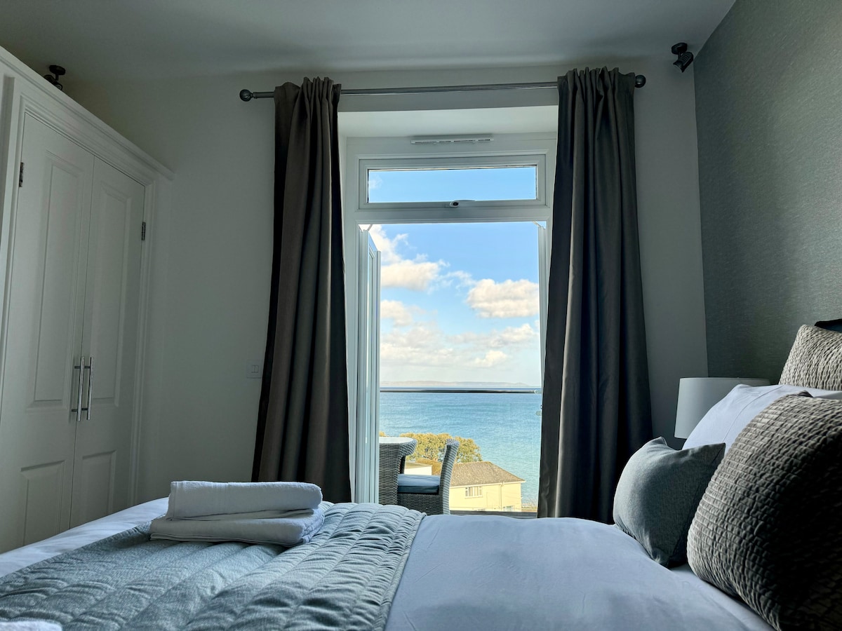 A master bedroom features a spacious bed adorned with a soft grey throw and rolled towels, positioned near a large window. Stunning ocean views are visible through the open window, framed by elegant curtains that enhance the atmosphere of serenity.