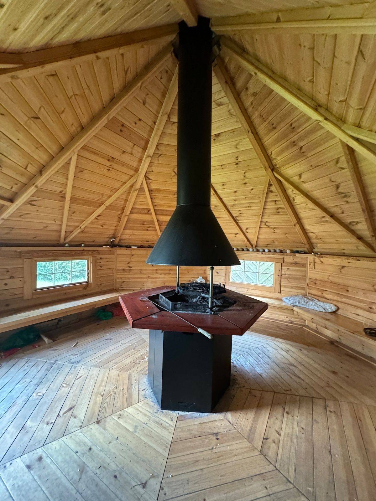An interior view of a wooden grill hut features a central fireplace with a black chimney. The space is surrounded by hexagonal wooden walls and illuminated by natural light filtering through multiple windows. A patterned wooden floor completes the rustic atmosphere.