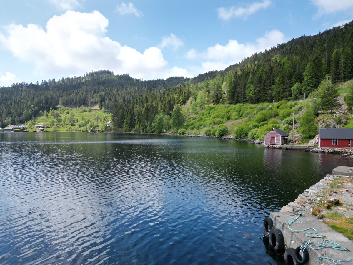 A serene lake is surrounded by lush green hills and dense forests, creating a peaceful natural setting. A small red boathouse is positioned at the water's edge, while a pier extends into the lake. The sky is partly cloudy, reflecting scenic beauty.